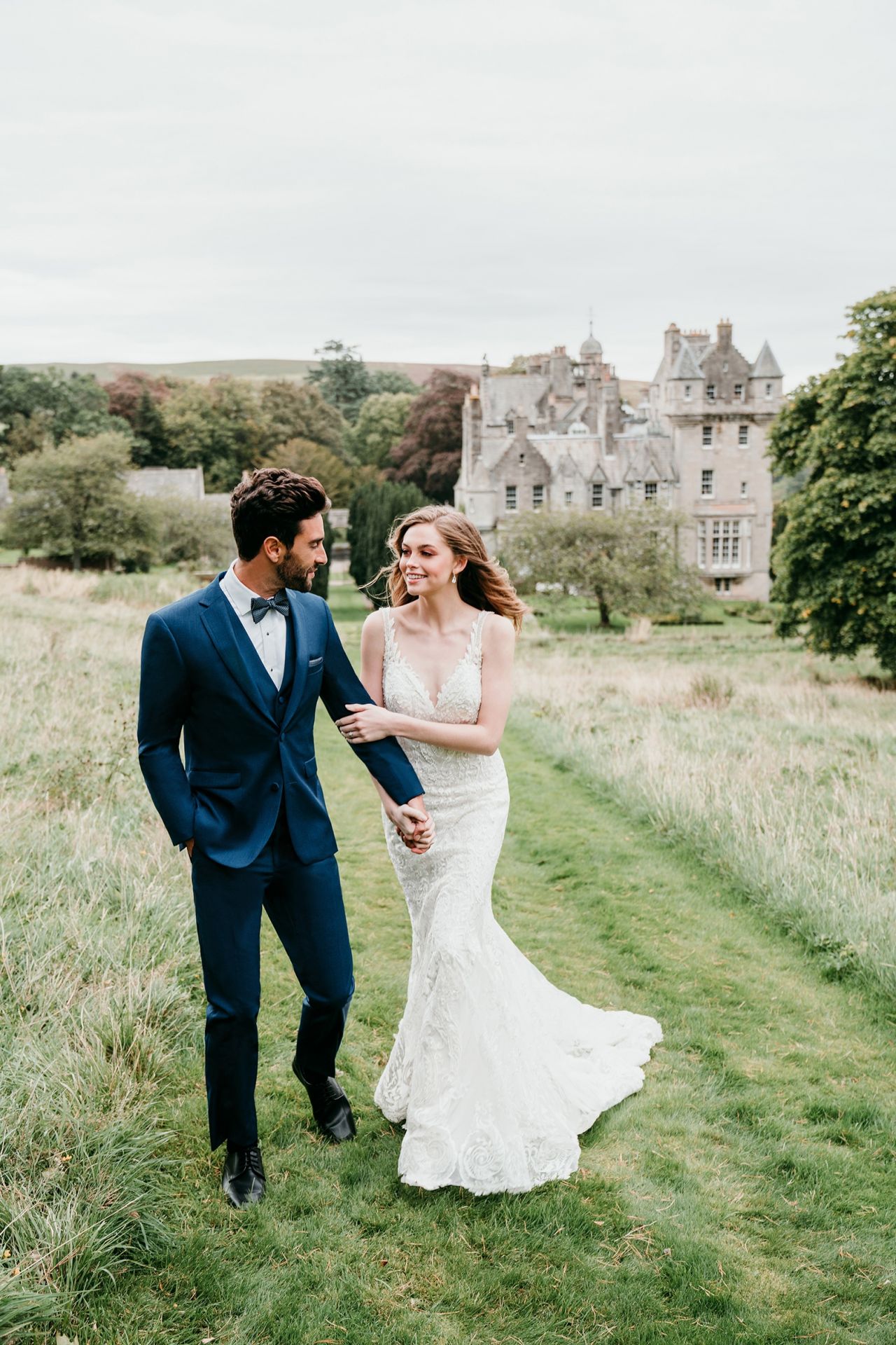 Groom wearing a dark blue tuxedo holding hands with bride who is wearing a strappy wedding dress