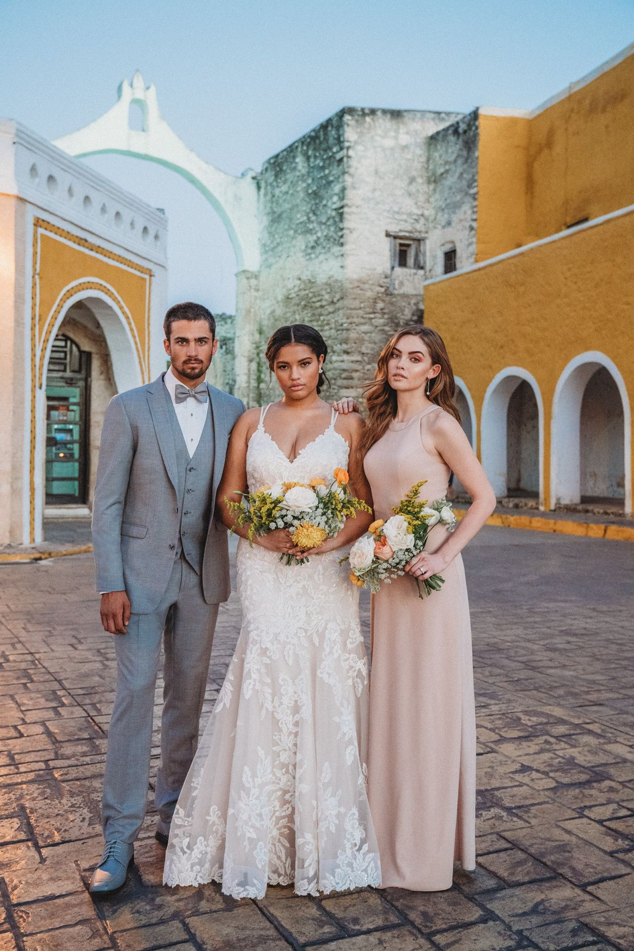Bride wearing a lace wedding dress with groom in a grey suit and maid of honor in soft blush bridesmaid dress